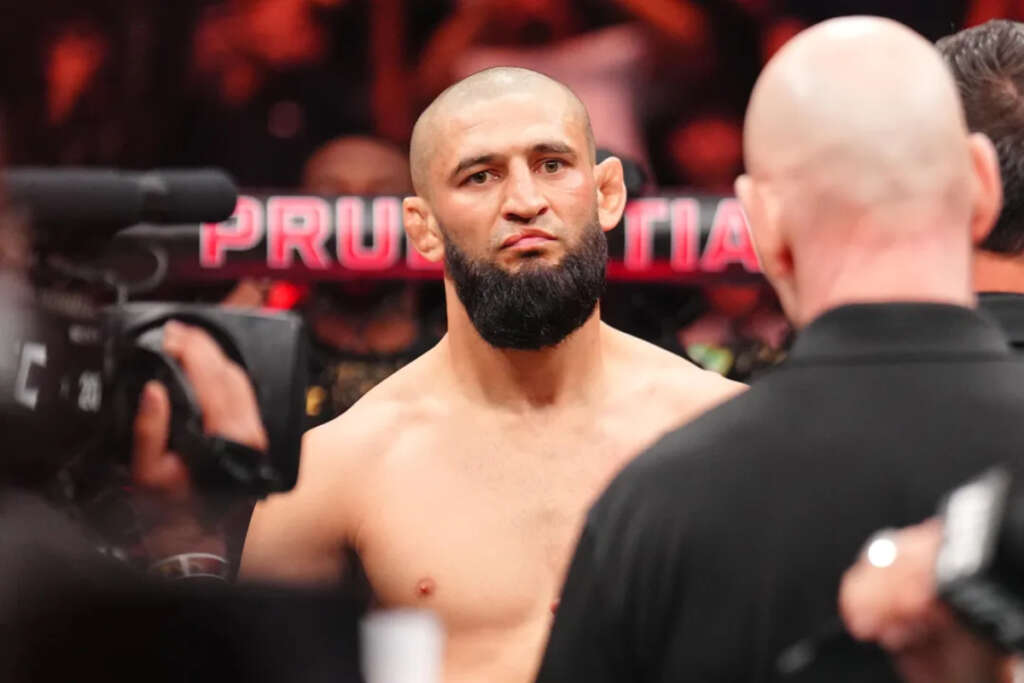 Shirtless male mixed martial artist with a beard stands in a ring, reporters and cameras nearby before a crowd under red lighting