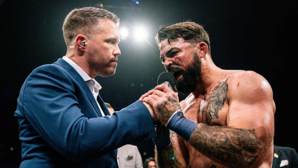 Interviewer in a blue suit holds a microphone to a shirtless, tattooed fighter with facial injuries in a boxing ring.