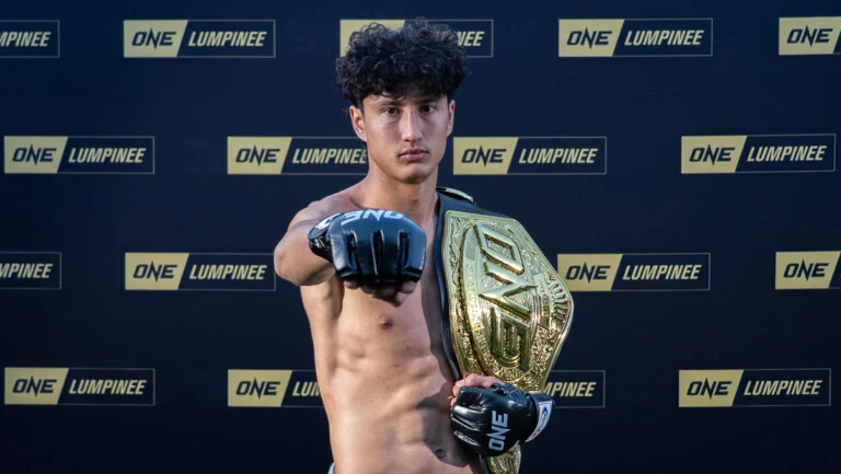 Young male MMA fighter, shirtless, extends fist toward the camera while holding a large championship belt; backdrop features ONE LUMPINEE logos.
