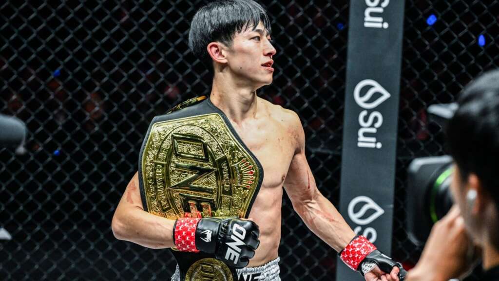MMA fighter standing in the cage with a gold championship belt draped over his shoulder, fists clenched and looking to the side.