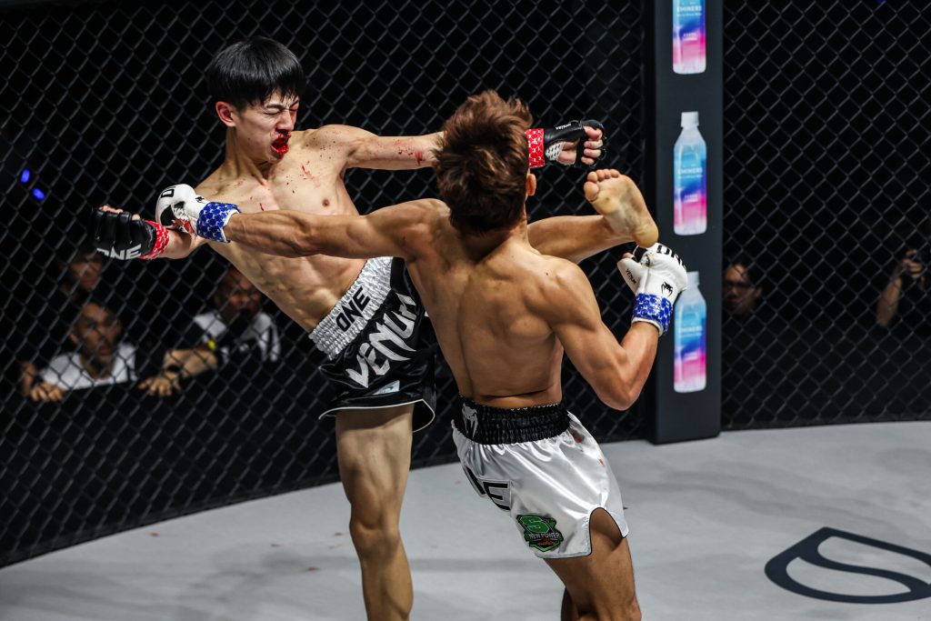 Two male mixed martial artists exchange a high kick inside a cage, one delivering a knee to the other’s midsection; spectators in the background.