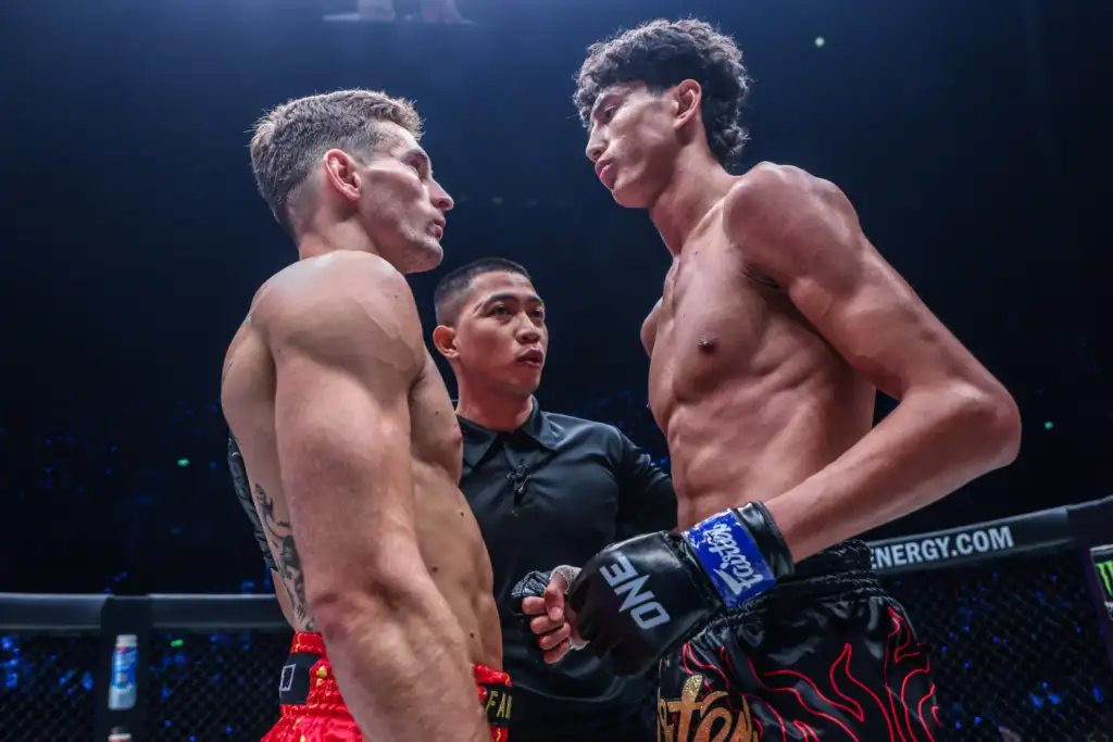 Two shirtless boxers face off in a ring, forehead-to-forehead, with a referee between them under arena lights.