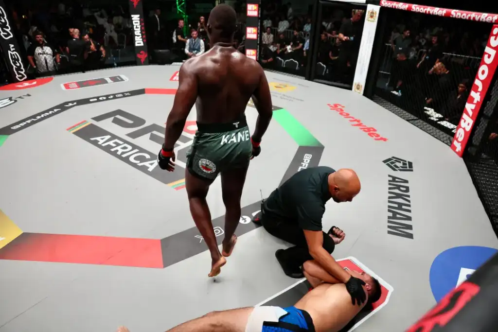MMA fighter in green shorts stands over a fallen opponent on the octagon mat as a referee kneels beside them, crowd watching.