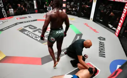 MMA fighter in green shorts stands over a fallen opponent on the octagon mat as a referee kneels beside them, crowd watching.