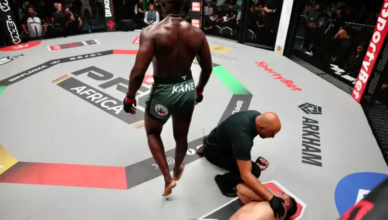 MMA fighter in green shorts stands over a fallen opponent on the octagon mat as a referee kneels beside them, crowd watching.