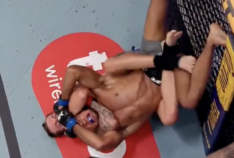Screenshot-2026-10-10-at-105649-PM - AfroCombat Two mixed martial artists grapple on a blue mat; the top fighter applies a choke while the bottom fighter braces near the cage, both wearing gloves.