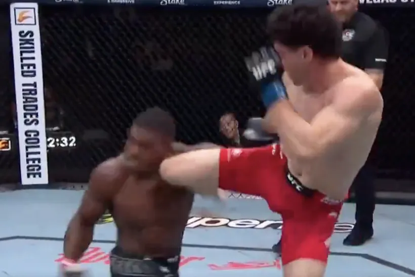 A fighter in red shorts delivers a high kick to a shirtless opponent inside a cage.