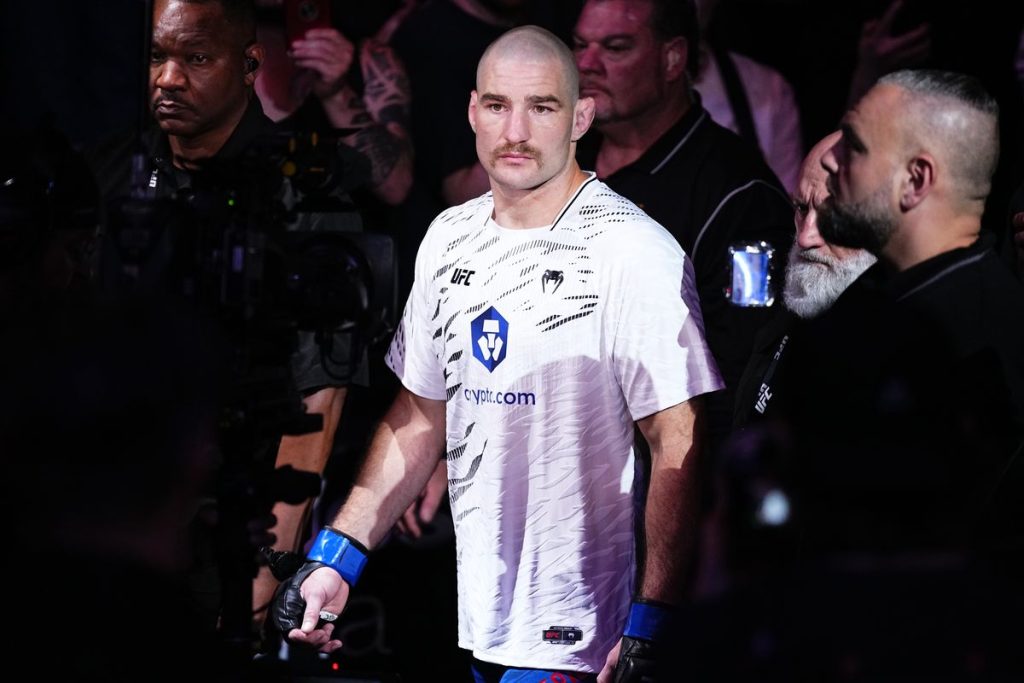 Male UFC fighter walks toward octagon with blue gloves, white shirt, focused expression amid photographers and crowd.