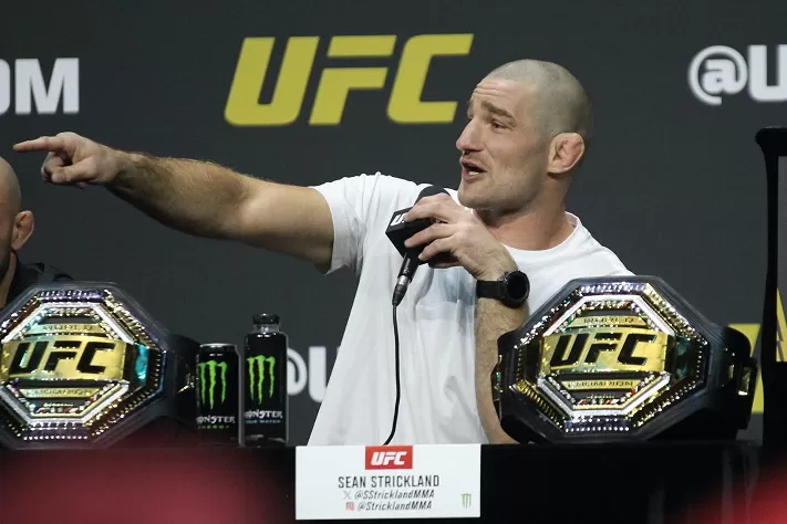 Sean Strickland speaks at a UFC press conference, pointing while holding a mic, with UFC belts visible on the table.