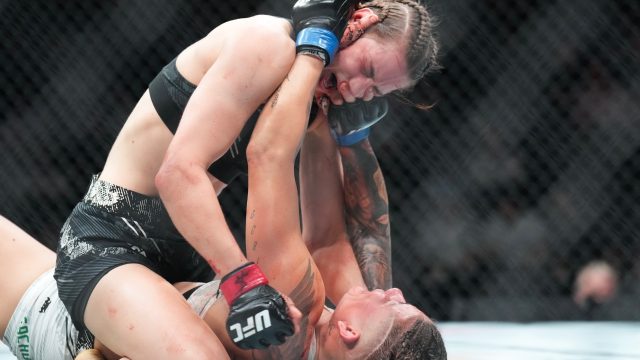 Female mixed martial artists grappling inside a cage; one on top in a ground-and-pound position, delivering strikes to the opponent's head with red gloves.