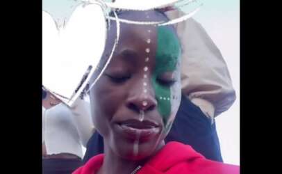 Close-up of a woman in a red hoodie, half her face painted green with white dot lines and a white overlay across the camera frame.