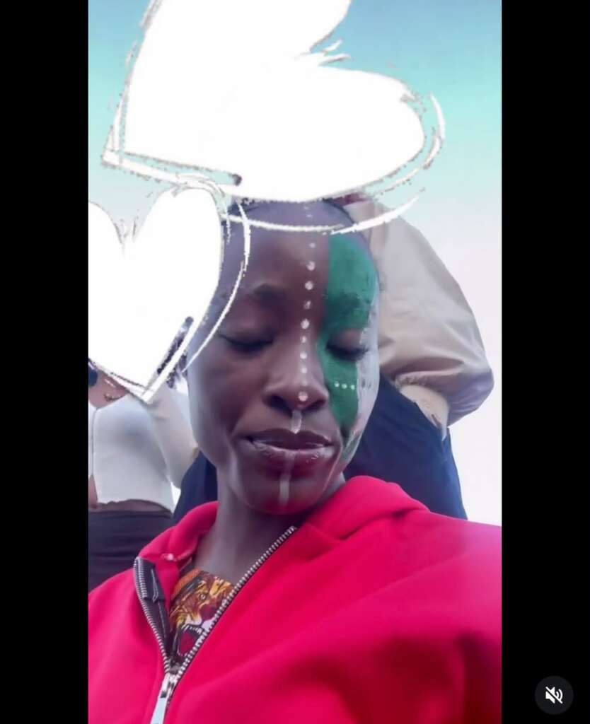 Close-up of a woman in a red hoodie, half her face painted green with white dot lines and a white overlay across the camera frame.
