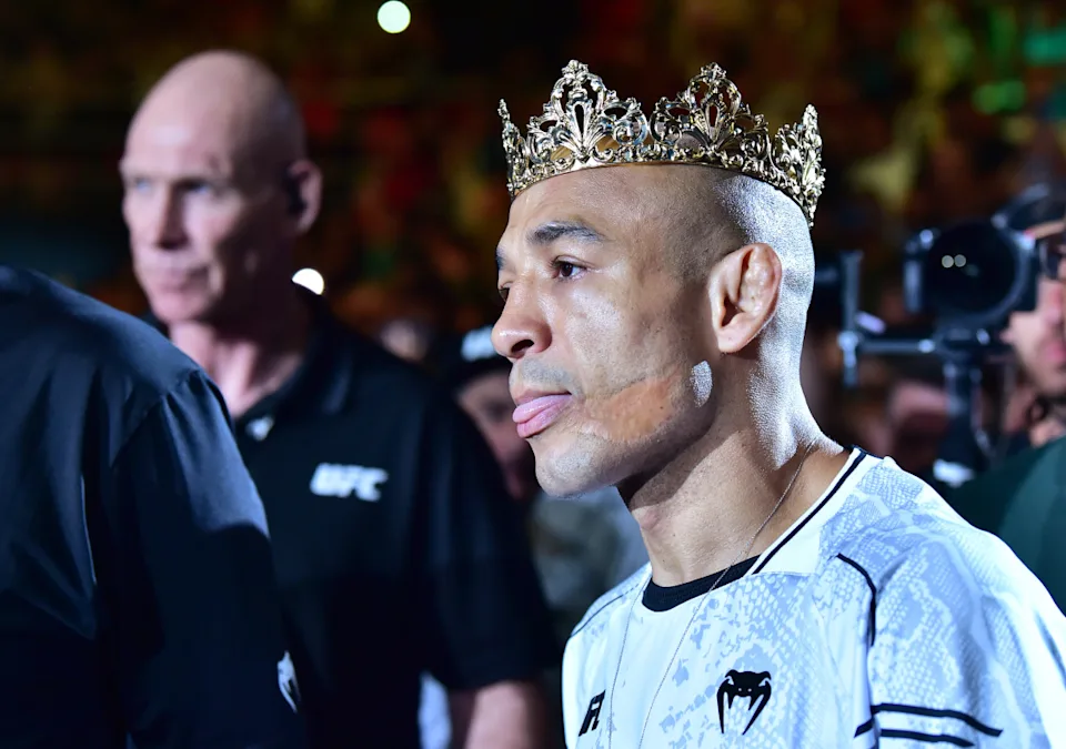 MMA fighter wearing a gold crown during a walkout, with photographers in the background at a crowded arena.