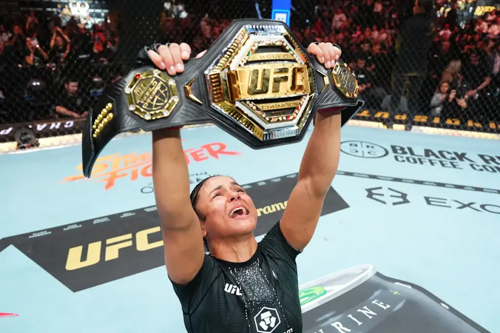 Female UFC fighter triumphs in the octagon, lifting a championship belt above her head as cheering crowd looks on.