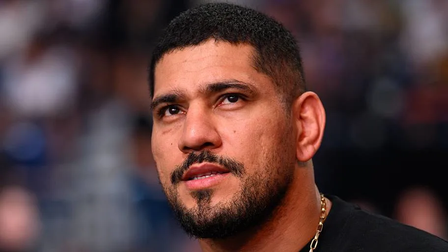 Close-up portrait of a man with short dark hair and a beard, wearing a gold chain, looking slightly upward.