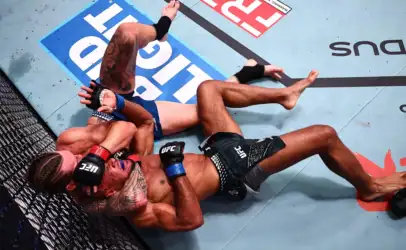 Two UFC fighters grapple on the mat inside the fenced octagon, one on top pinning the other during a ground exchange.