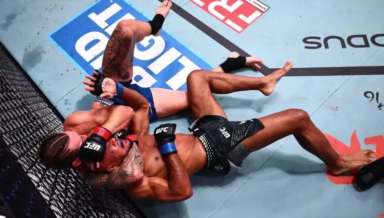 Two UFC fighters grapple on the mat inside the fenced octagon, one on top pinning the other during a ground exchange.