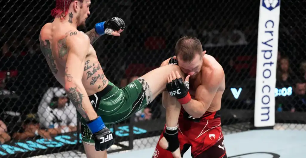 Two UFC fighters clash in the cage: one delivering a high kick while the other braces and blocks with his arms.