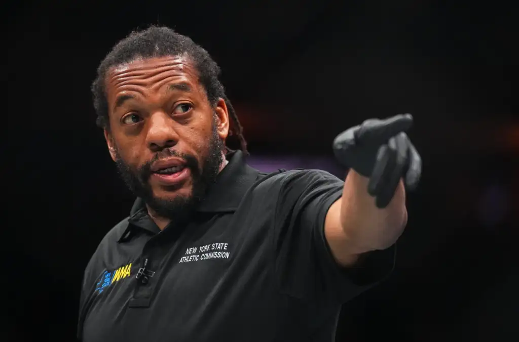 Man in a black polo with NYS Athletic Commission logo points toward the camera with a gloved hand on a dark stage/background.