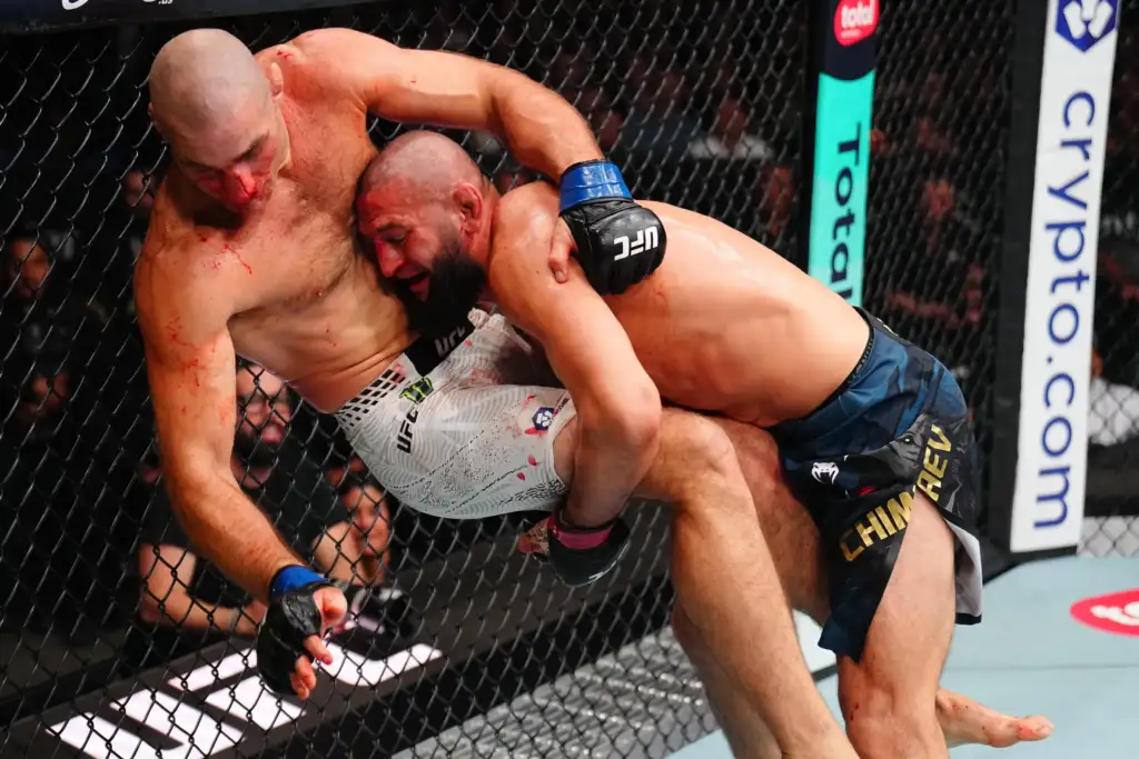 Two UFC fighters grapple against a chain-link cage, one with blue gloves leaning in with a bloody face and the other in white shorts wrapped around him.