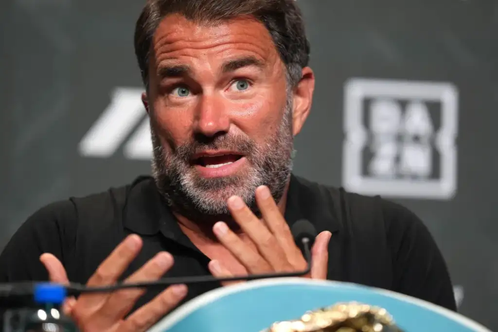 Bearded man at a press conference speaking into a microphone, gesturing with his hands in front of a backdrop