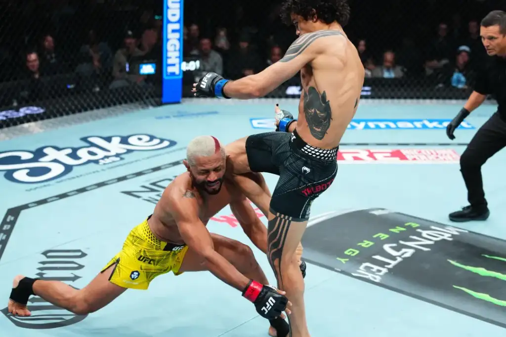 Two UFC fighters clash in the octagon: a shirtless fighter in black shorts delivers a knee as his opponent in yellow trunks struggles on the mat.