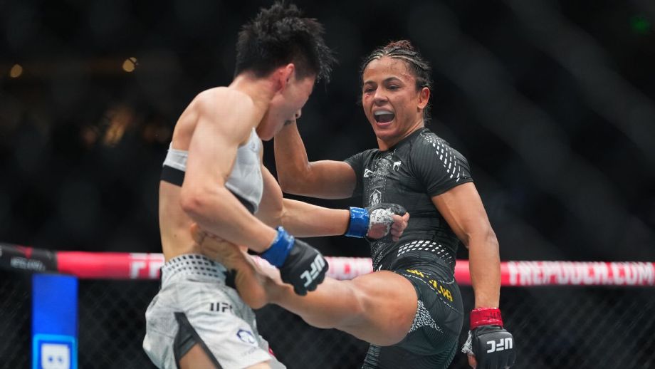 Two mixed martial artists clash inside the UFC octagon as the woman in black attacks with a high kick toward the opponent wearing white shorts with blue gloves in the foreground.