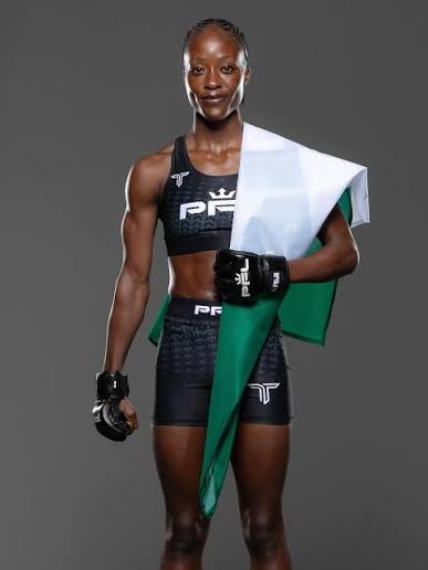 Female mixed martial artist standing in a studio, wearing black fight shorts and gloves, holding a green‑white‑red flag over her shoulder.