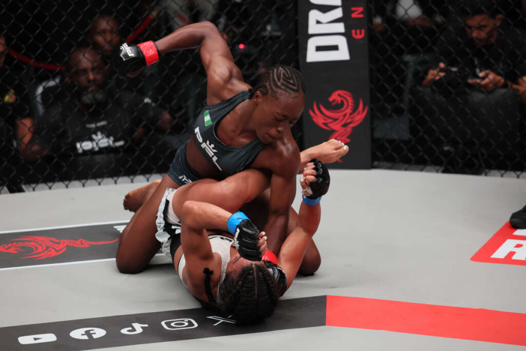 Two female MMA fighters grapple on the mat inside a cage; one on top delivering a control hold to the other on her back.