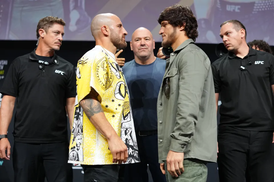 l-r-opponents-alexander-volkanovski-1115211840 - AfroCombat Two UFC fighters stare each other down on stage before a fight, flanked by staff and a backdrop in the distance.