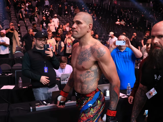 Bald, tattooed male fighter in colorful shorts and red wrist wraps walks through a crowded arena as photographers and fans take pictures nearby.
