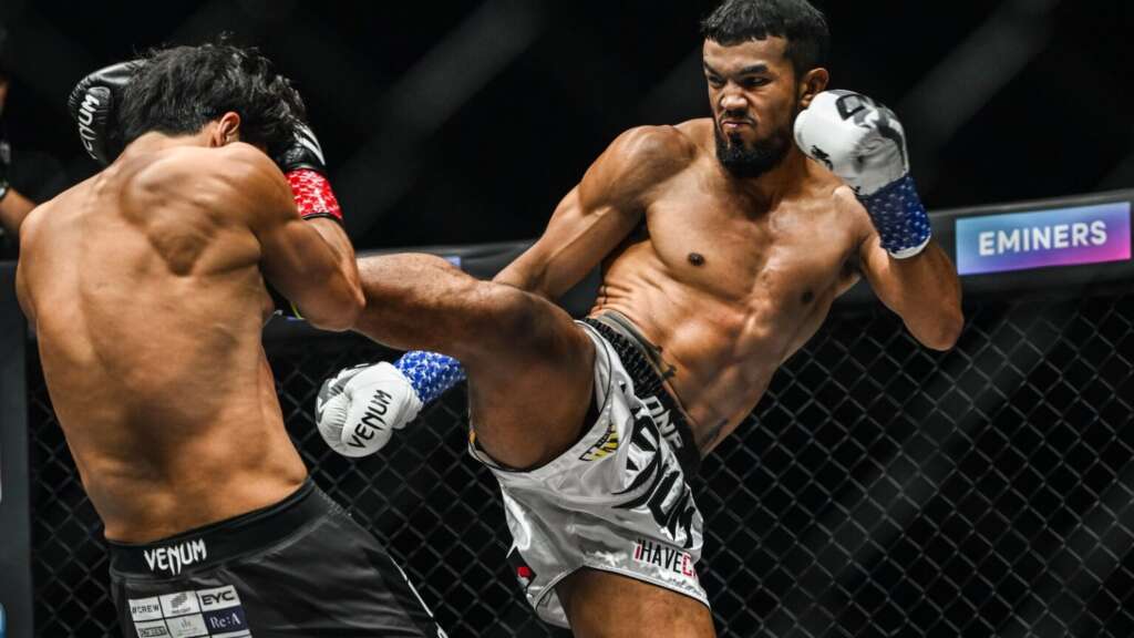 Two shirtless fighters in a cage exchange a high kick; one thows a powerful kick toward the other's torso while both guard their heads. Round-house action in a martial arts match.